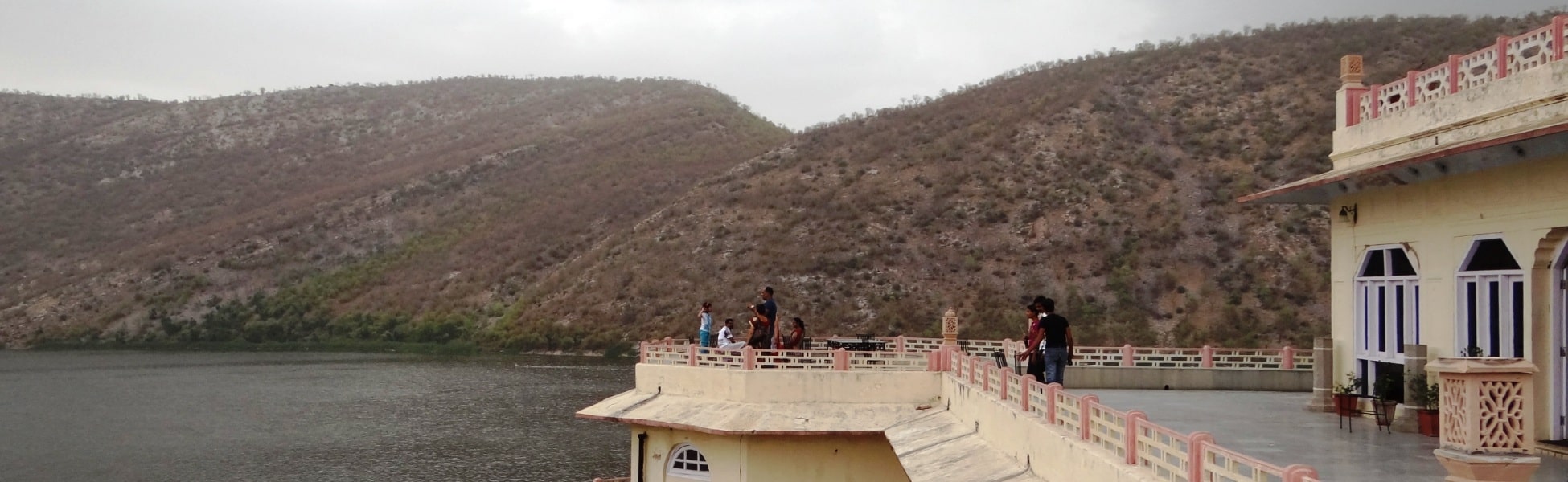Tourists standing on the cream-colored ornate terrace of Siliserh Lake Palace in Alwar, overlooking the calm Siliserh Lake with the dry, rugged Aravalli Hills in the background under an overcast sky. fixer in Balrampur, film fixer in Balrampur, media fixer in Balrampur, movie fixer in Balrampur, production fixer in Balrampur, line producer in Balrampur
