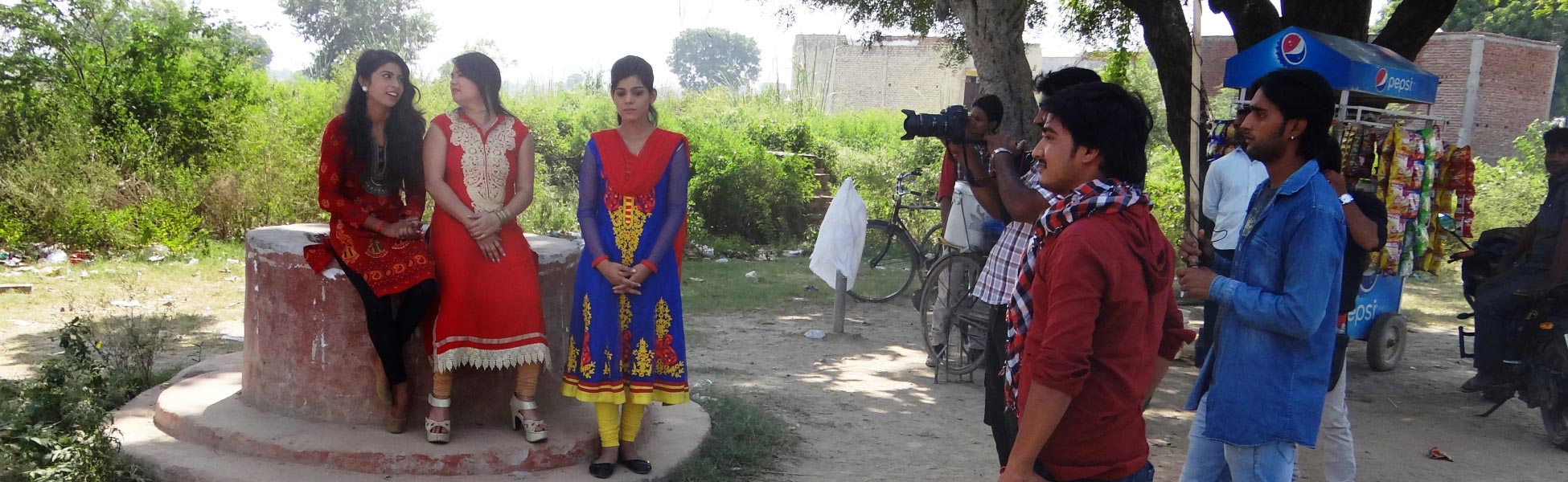 Behind-the-scenes photo of an indie Indian film crew filming 'Don't Harass the Girl' in Ghaziabad, Uttar Pradesh. Three actresses in traditional attire sit by a concrete well while a camera operator and crew frame the shot. film production house in Bangalore South, video production house in Bangalore South, tv production house in Bangalore South, production house in Bangalore South, film production company in Bangalore South, video production company in Bangalore South, tv production company in Bangalore South, production company in Bangalore South