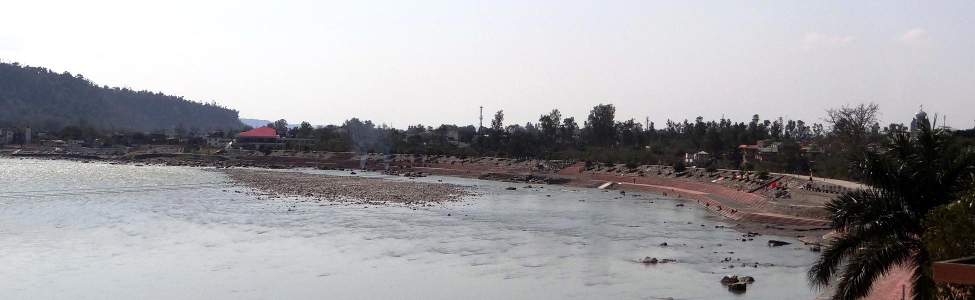Wide panoramic view of a scenic riverfront film shooting location in India, featuring a sweeping curved concrete ghat embankment, a shallow rocky riverbed, and lush green hills with a red-roofed building in the background. fixer in Bangalore Urban, film fixer in Bangalore Urban, media fixer in Bangalore Urban, movie fixer in Bangalore Urban, production fixer in Bangalore Urban, line producer in Bangalore Urban