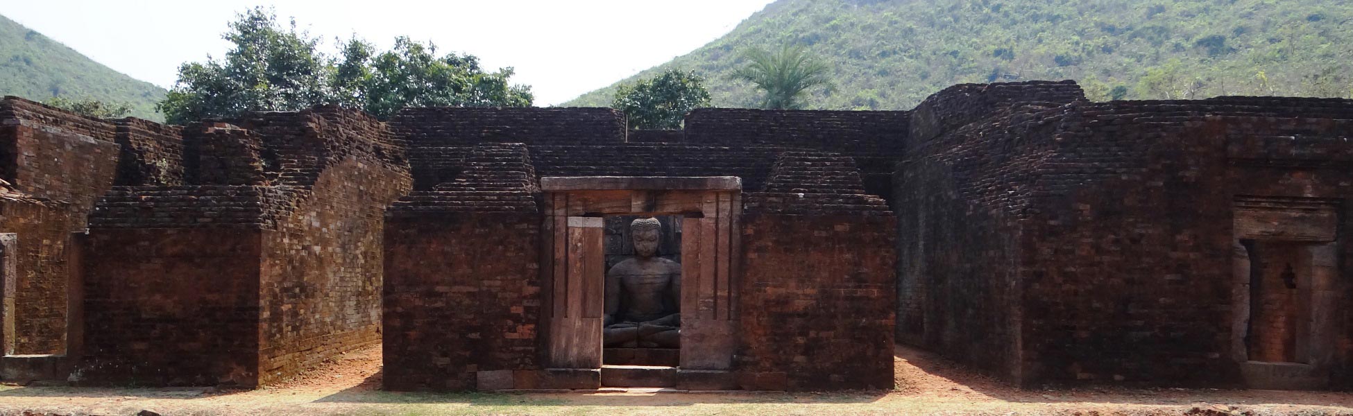 Ancient stone seated Buddha statue visible through a brick shrine doorway at the ruins of Monastery 1 in the Udayagiri Buddhist Complex, Jajpur, Odisha, India, with a lush green hill in the background. film shooting locations in Bareilly, video shooting locations in Bareilly, tv shooting locations in Bareilly, shooting locations in Bareilly