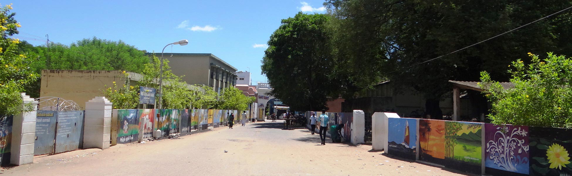 A wide-angle street view leading toward the Thanjavur Royal Palace complex from file 135.jpg, featuring vibrant cultural wall murals, lush green overhanging trees, and pedestrians under a bright blue sky. film shooting locations in Begusarai, video shooting locations in Begusarai, tv shooting locations in Begusarai, shooting locations in Begusarai