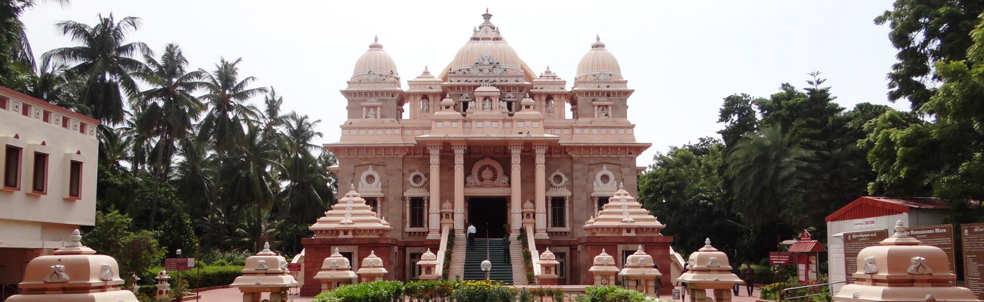 A wide front panoramic view of the grand pale-pink Universal Temple at Sri Ramakrishna Math in Mylapore, Chennai. The image captures its distinct multi-tiered architecture that blends Hindu Dravidian styles, Buddhist motifs, and Western structural elements, set against a backdrop of lush palm trees and manicured temple pathways. fixer in Belagavi, film fixer in Belagavi, media fixer in Belagavi, movie fixer in Belagavi, production fixer in Belagavi, line producer in Belagavi
