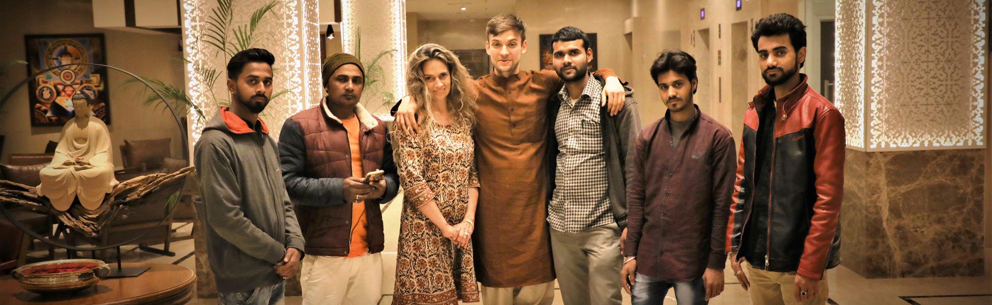 A group photo taken in a brightly lit hotel lobby features eight people: the professional camera crew from Trisna Films®, India, and their US client for a music video production in Bodhgaya, Bihar. They pose near a statue of a seated Buddha and decorative, illuminated pillars at the Lemon Tree Premier in Patna. film production house in india, film production house in Delhi, film production house in mumbai, film production house in pune, film production house in sonipat, film production house in begusarai, video production house in india, video production house in Delhi, video production house in mumbai, video production house in pune, video production house in sonipat, video production house in begusarai, tv production house in india, tv production house in delhi, tv production house in mumbai, tv production house in pune, tv production house in sonipat, tv production house in begusarai, production house in india, production house in delhi, production house in mumbai, production house in pune, production house in sonipat, production house in begusarai, film production company in india, film production company in delhi, film production company in mumbai, film production company in pune, film production company in sonipat, film production company in begusarai, video production company in india, video production company in delhi, video production company in mumbai, video production company in pune, video production company in sonipat, video production company in begusarai, tv production company in india, tv production company in delhi, tv production company in mumbai, tv production company in pune, tv production company in sonipat, tv production company in begusarai, production company in india, production company in delhi, production company in mumbai, production company in pune, production company in sonipat, production company in begusarai