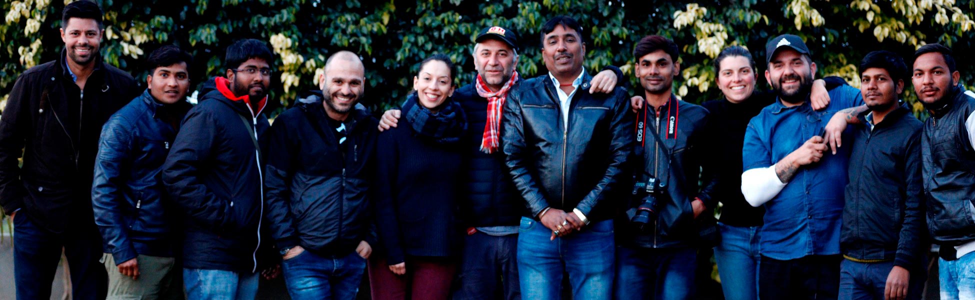 Group portrait of the diverse international film crew from Trisna Films®, the US, and Italy. The crew members are smiling and posing together outdoors in front of a leafy green background while on location in Ludhiana, Punjab, India, for the production of the web series 'All in The Family - Under One Roof'. film production house in india, video production house in india, tv production house in india, production house in india, film production company in india, video production company in india, tv production company in india, production company in india