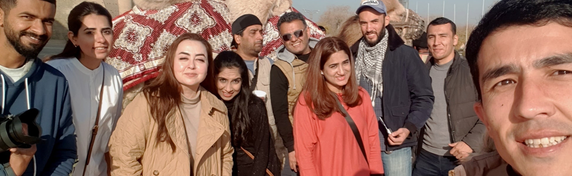 The Trisna Films® crew and the Maria.B. team taking a group selfie with decorated camels at the ancient Ark of Bukhara fortress in Uzbekistan during a professional fashion ad production. filmmaking workshop in india, cinematography workshop in india, camera workshop in india, editing workshop in india, vfx workshop in india, animation workshop in india, photography workshop in india, direction workshop in india, script writing workshop in india