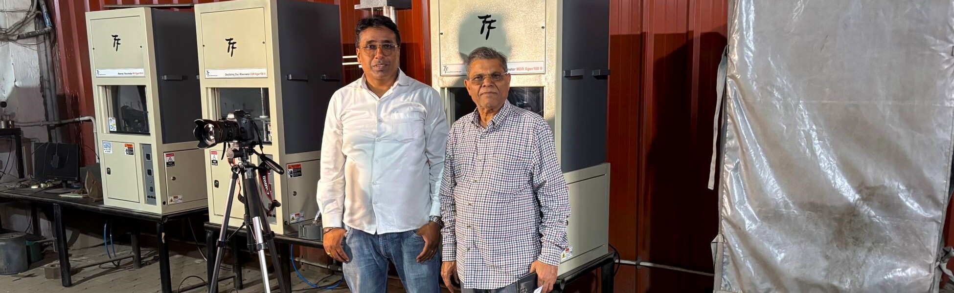 Behind-the-scenes of Trisna Films® crew during an industrial video production at a manufacturing facility in Kundli, Sonipat, Haryana. The image features two professional crew members standing alongside specialized industrial testing machinery, with a DSLR camera mounted on a tripod in the foreground, showcasing on-location expertise in industrial cinematography. film shooting locations in india, video shooting locations in india, tv shooting locations in india, shooting locations in india