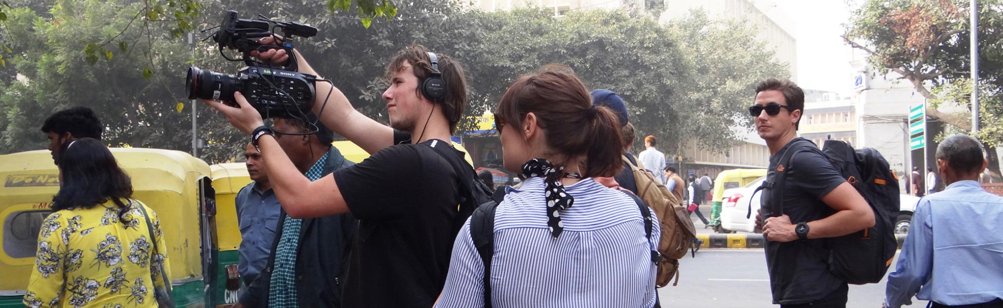 A British production crew from Insight TV filming on a bustling street in New Delhi, India. A cameraman holds a professional video camera, a crew member wears audio headphones, and iconic yellow-and-green auto-rickshaws are visible in the background. fixer in Bageshwar, film fixer in Bageshwar, media fixer in Bageshwar, movie fixer in Bageshwar, production fixer in Bageshwar, line producer in Bageshwar