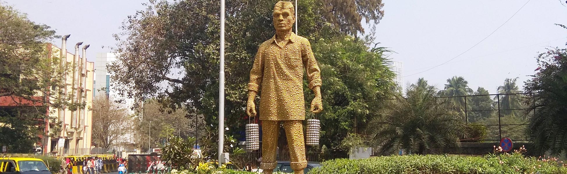 A wide daytime shot of the 13-foot-tall golden-colored stainless steel disc statue of a Mumbai Dabbawala wearing a traditional Gandhi topi and holding tiffin carriers, located at the Haji Ali junction roundabout on Tardeo Road in Mumbai, Maharashtra, India. fixer in Balaghat, film fixer in balaghat, media fixer in balaghat, movie fixer in balaghat, production fixer in balaghat, line producer in Balaghat